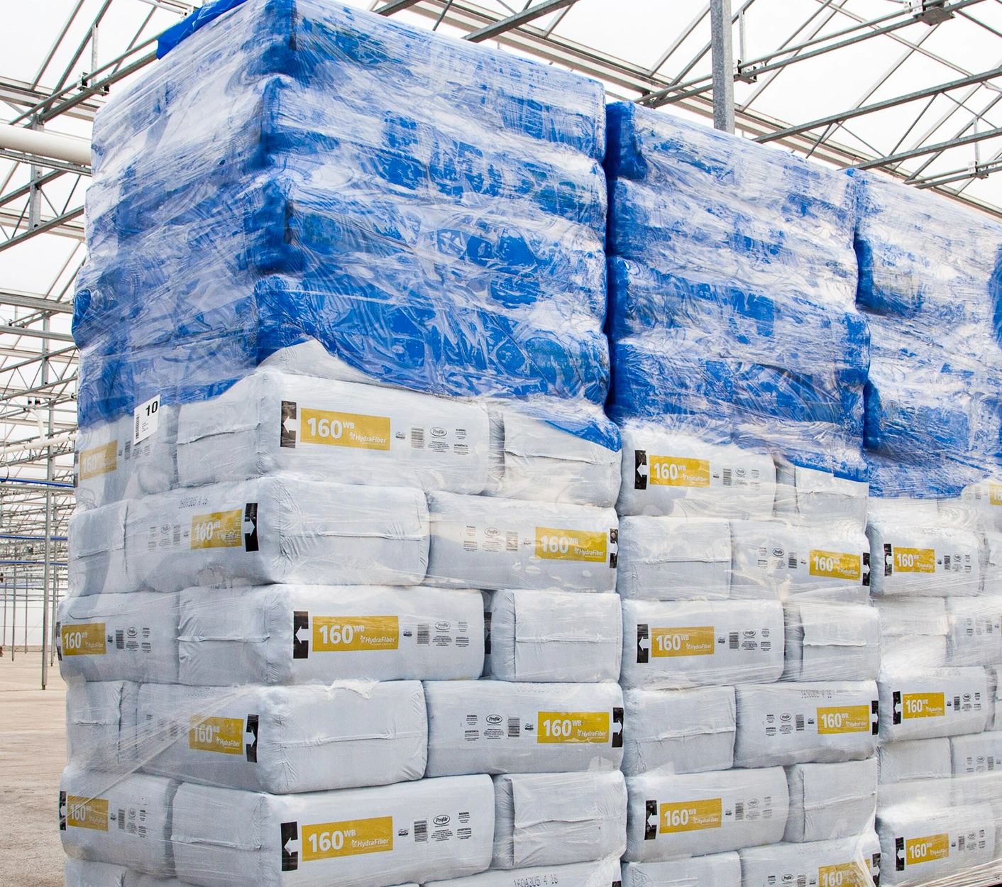 Stacks of white bags wrapped in blue plastic are piled on pallets inside a large warehouse or industrial facility with a glass ceiling.