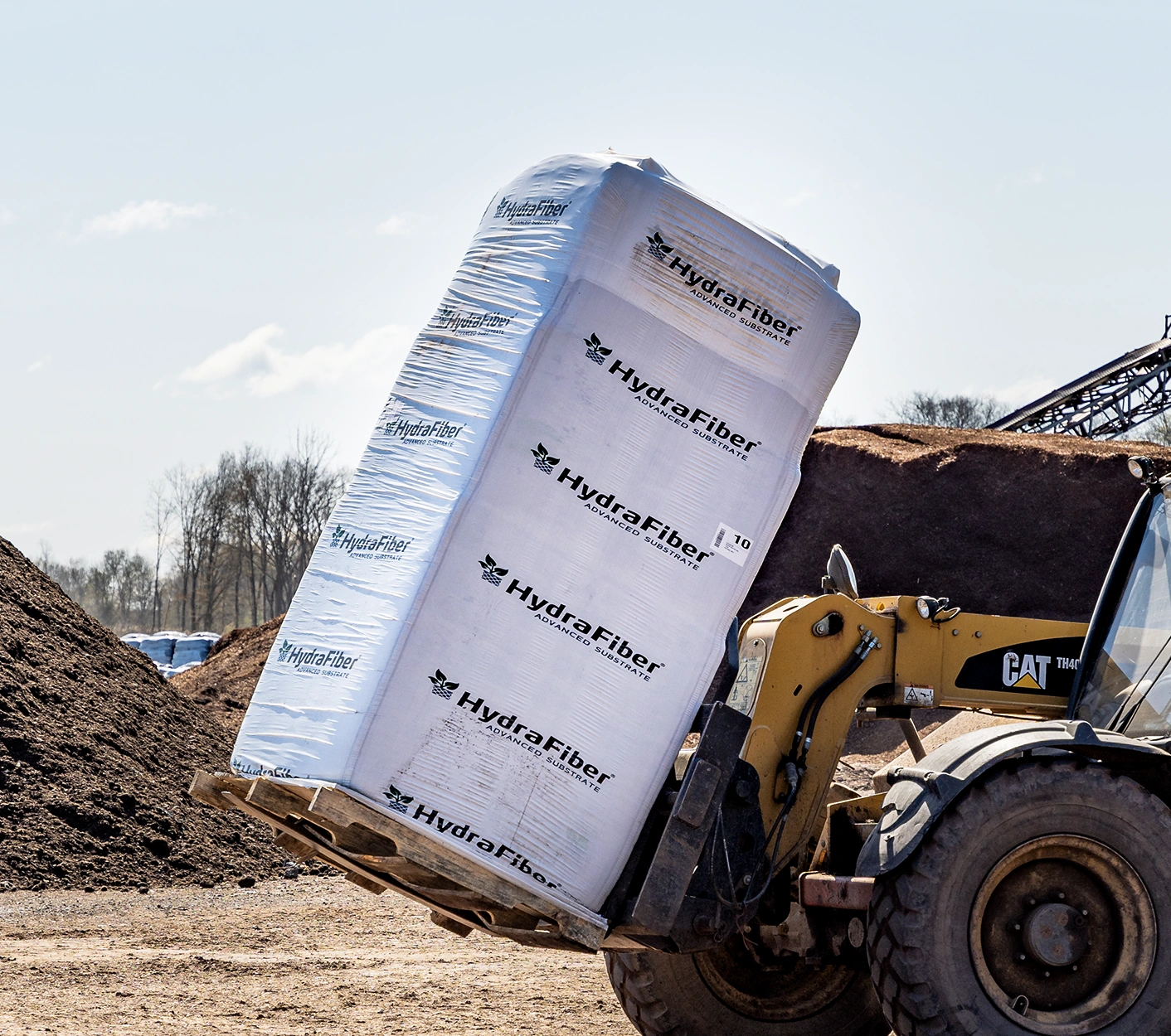 A forklift moves a large, wrapped pallet labeled HydraFiber at an outdoor industrial site, with dirt piles and trees visible in the background.