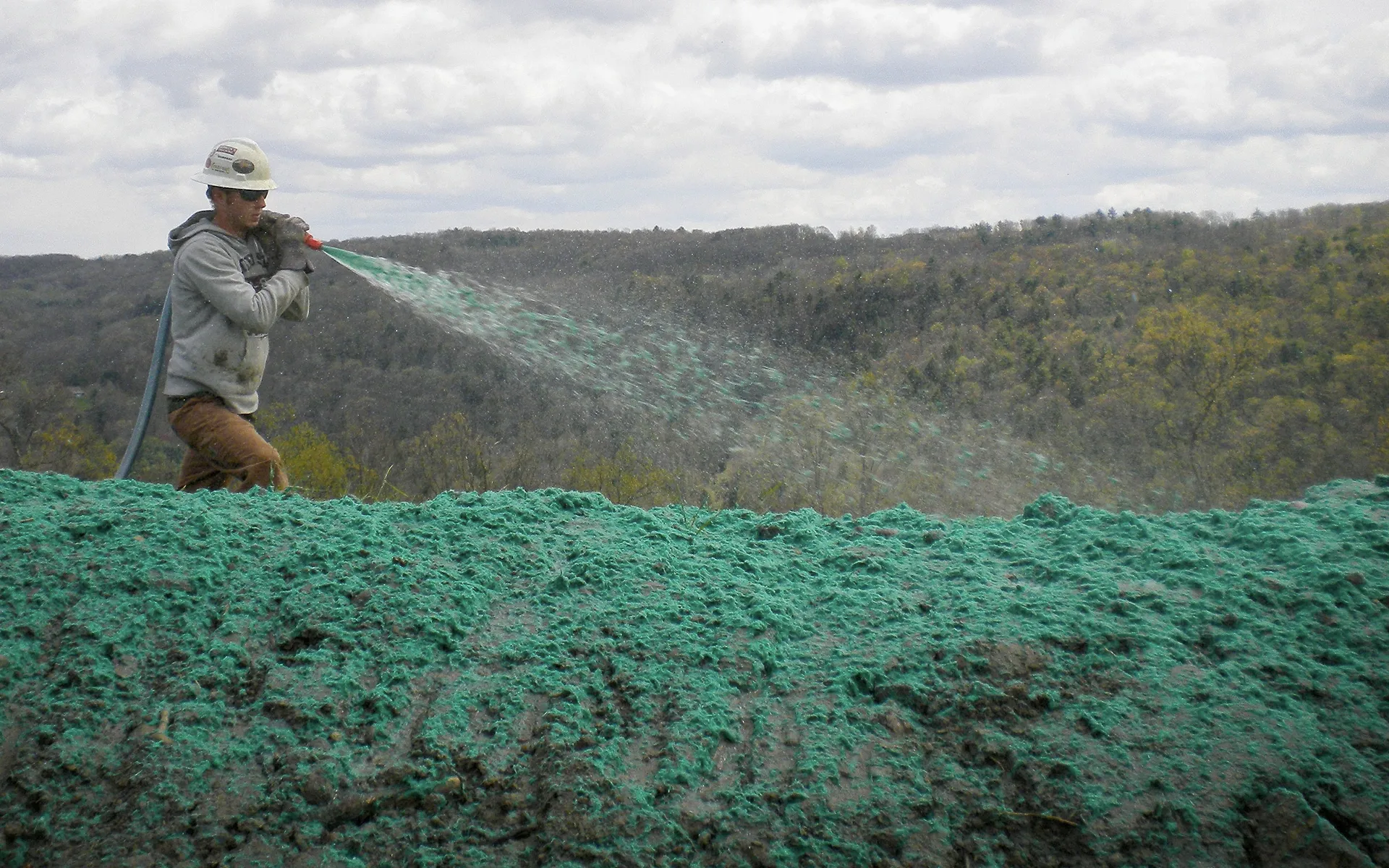 A worker in a hard hat sprays green hydroseed mixture onto a bare slope, with a forested landscape and cloudy sky in the background.