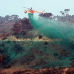 An orange helicopter drops green fire retardant over a hillside, with scattered trees and vegetation below, helping prevent wildfire spread.