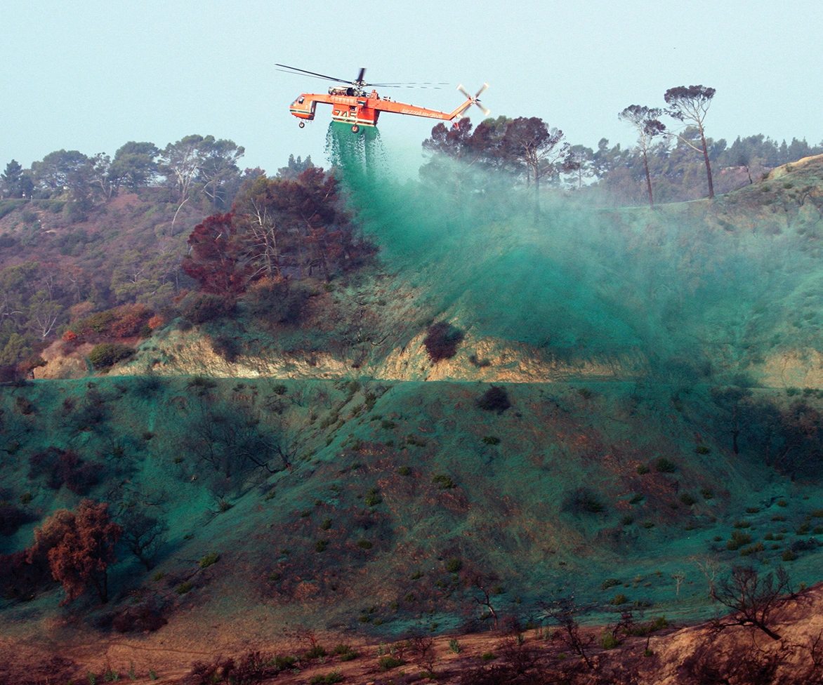 An orange helicopter drops green fire retardant over a hillside, with scattered trees and vegetation below, helping prevent wildfire spread.