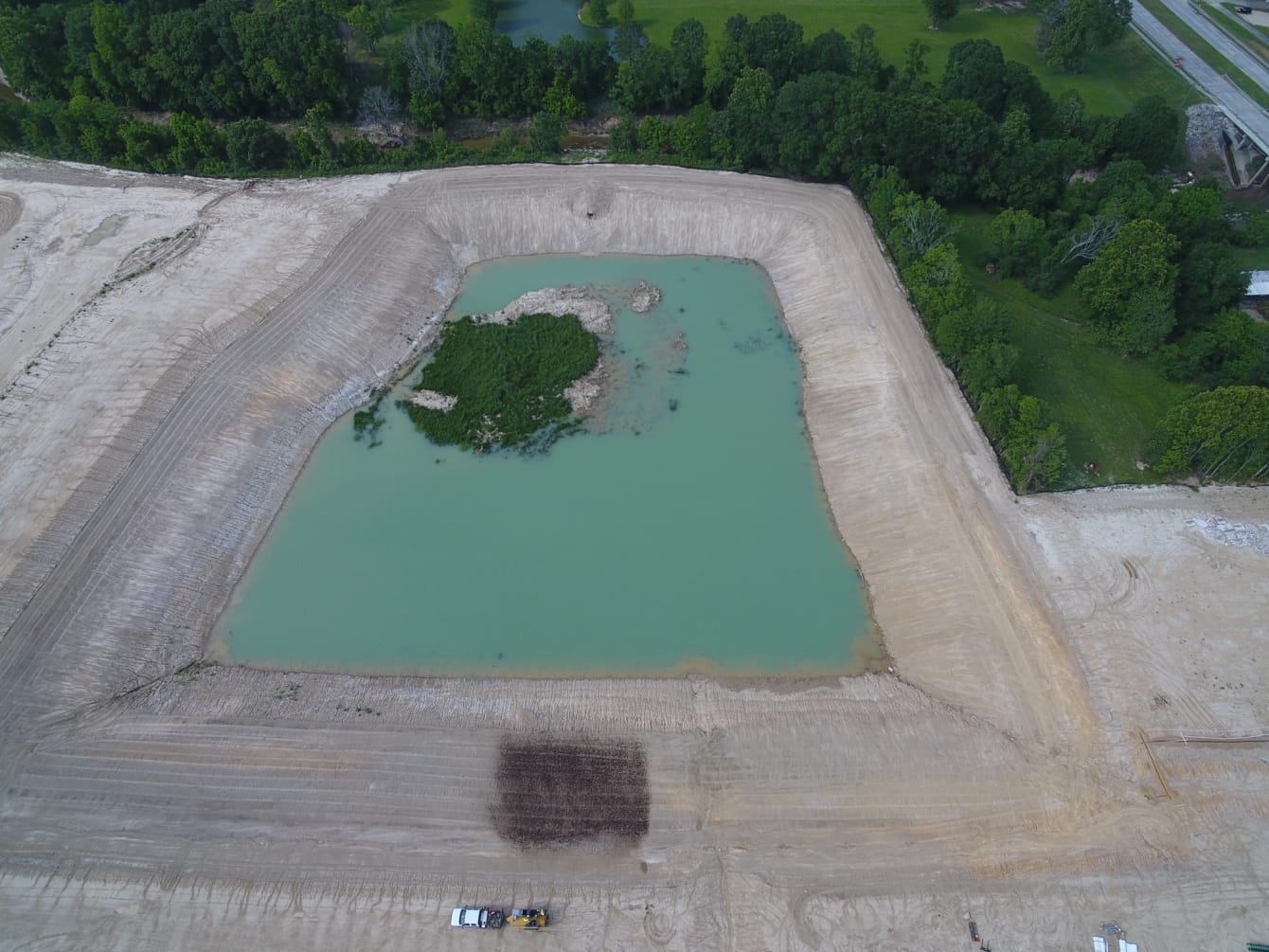 Aerial view of a large rectangular excavation filled with water and small patches of vegetation in the center, surrounded by dirt, with trees and grassy areas in the background.