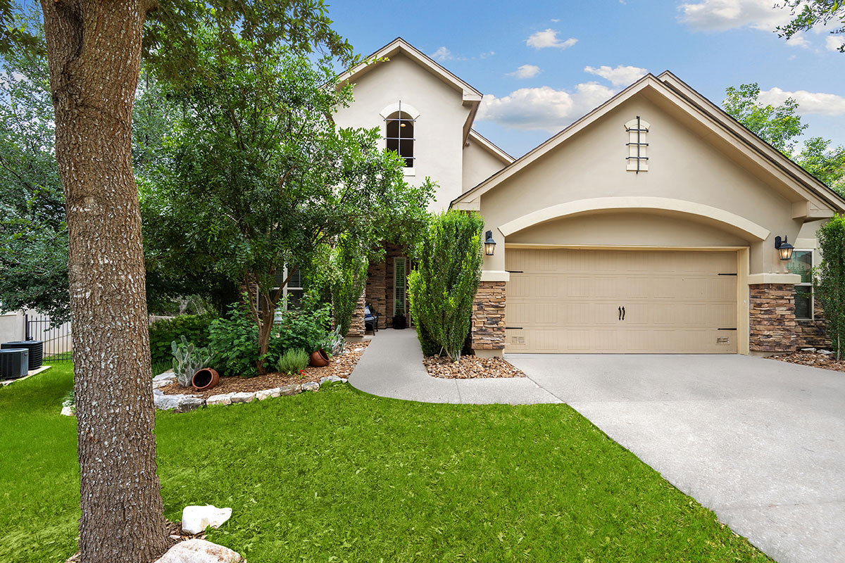 Two-story beige house with a double garage, arched entryway, and stone accents. Green lawn, trees, and shrubs surround the walkway to the front door. Blue sky with scattered clouds above.