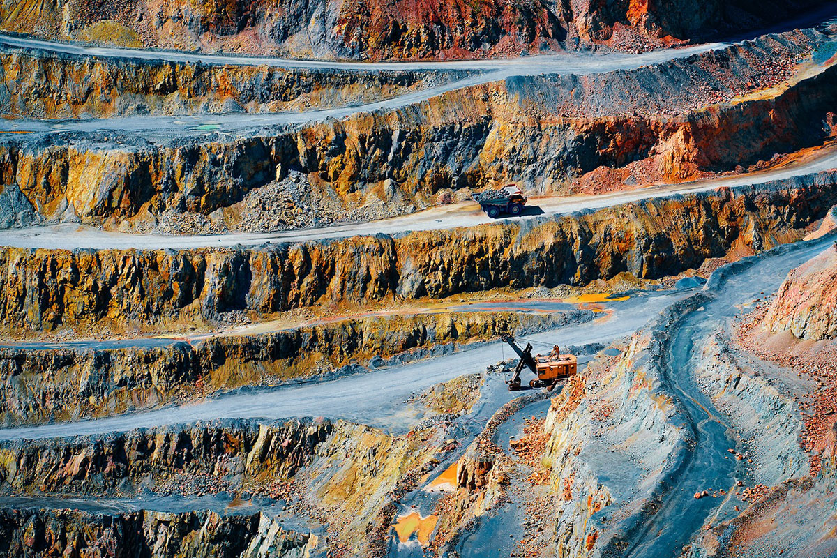 A large open-pit mine with terraced, rocky layers. Two trucks and an excavator are visible on the winding roads, highlighting the scale and industrial activity in the colorful, mineral-rich landscape.
