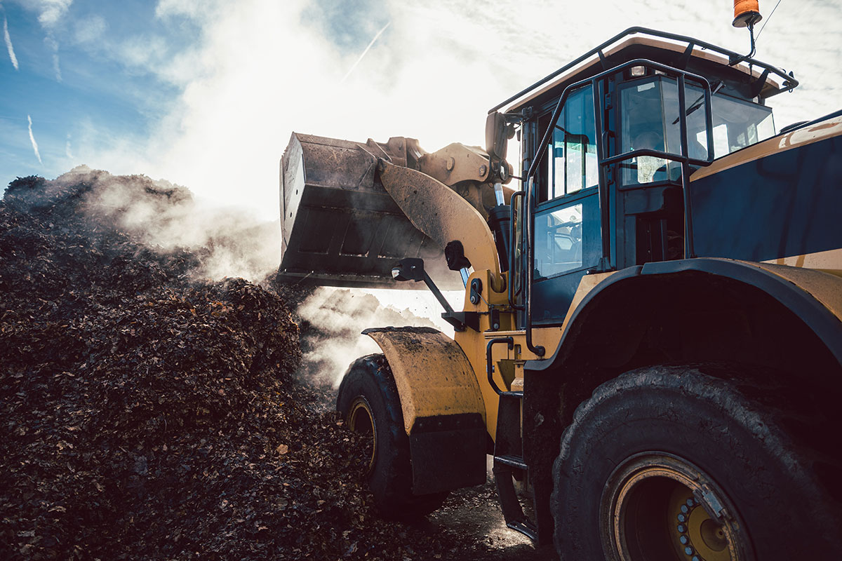 A yellow bulldozer scoops a large pile of dirt and debris, with steam or dust rising around it under a partly cloudy sky.