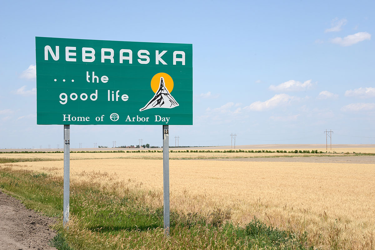A green road sign reads Nebraska... the good life. Home of Arbor Day by a golden wheat field under a blue sky with scattered clouds.