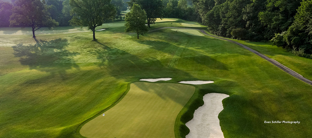 Aerial view of a lush golf course with a green, sand traps, trees, and winding paths, surrounded by dense forest under soft sunlight.