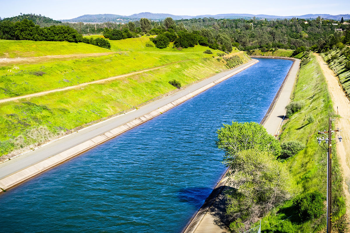 A wide, blue canal runs through grassy, green hills under a clear sky, bordered by concrete embankments and a dirt road with power lines on one side.