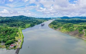 A large cargo ship travels through a wide canal surrounded by lush green hills under a partly cloudy sky.