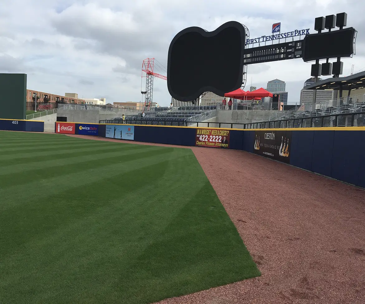 Professional baseball field with neatly maintained grass and warning track, with stadium seating and scoreboard in the background.