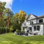 Large white house with a landscaped backyard lawn, patio seating area, and trees with autumn foliage.