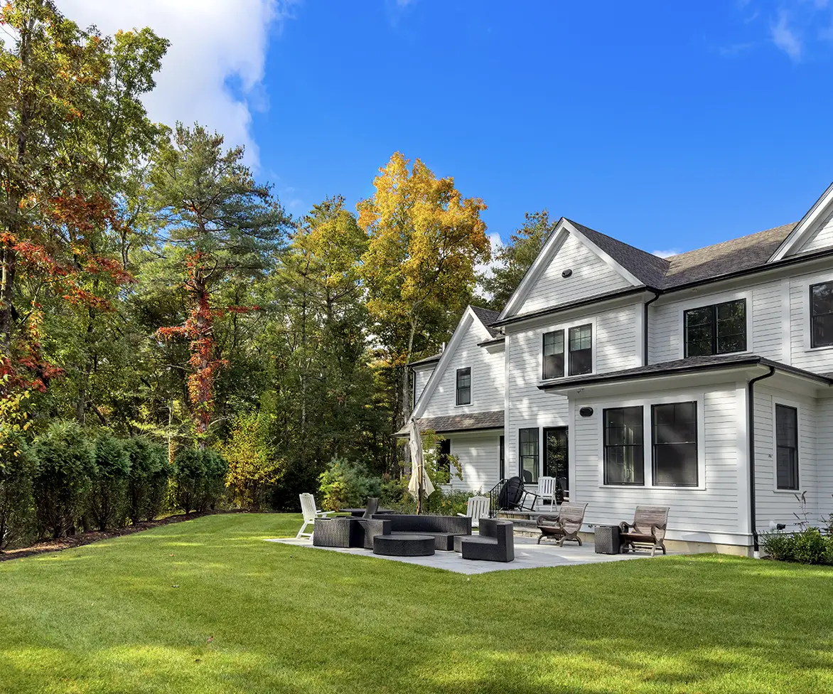 Large white house with a landscaped backyard lawn, patio seating area, and trees with autumn foliage.