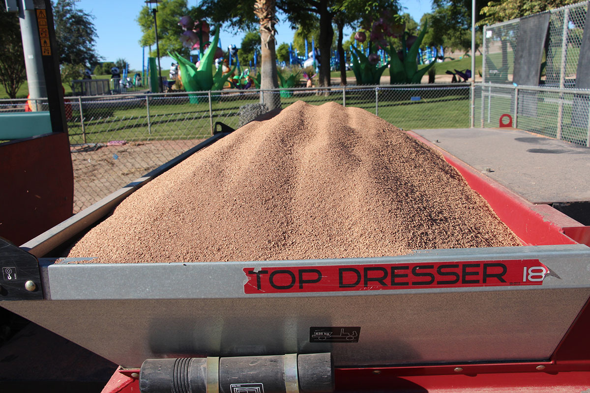A red Top Dresser 18 machine holds a load of reddish-brown sand or soil, with a park, trees, and playground equipment visible in the background.