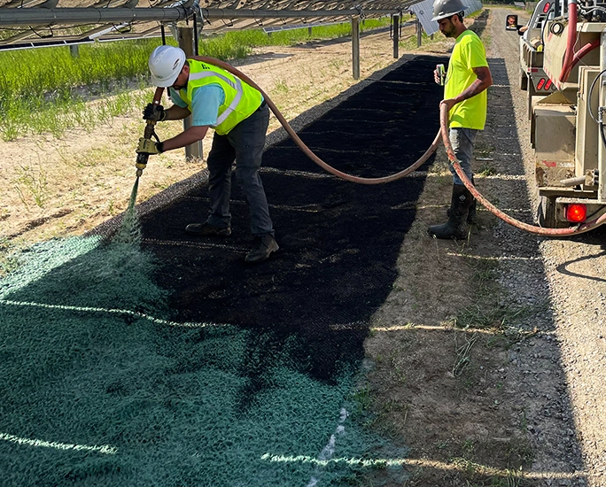 Two workers in safety gear spray green hydroseed onto soil under a metal structure, using a hose connected to a tank, to promote grass growth in a sunlit outdoor area.