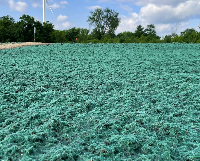 A field covered with bright green hydroseed mulch, used for planting grass, with trees, a wind turbine, and a partly cloudy sky in the background.