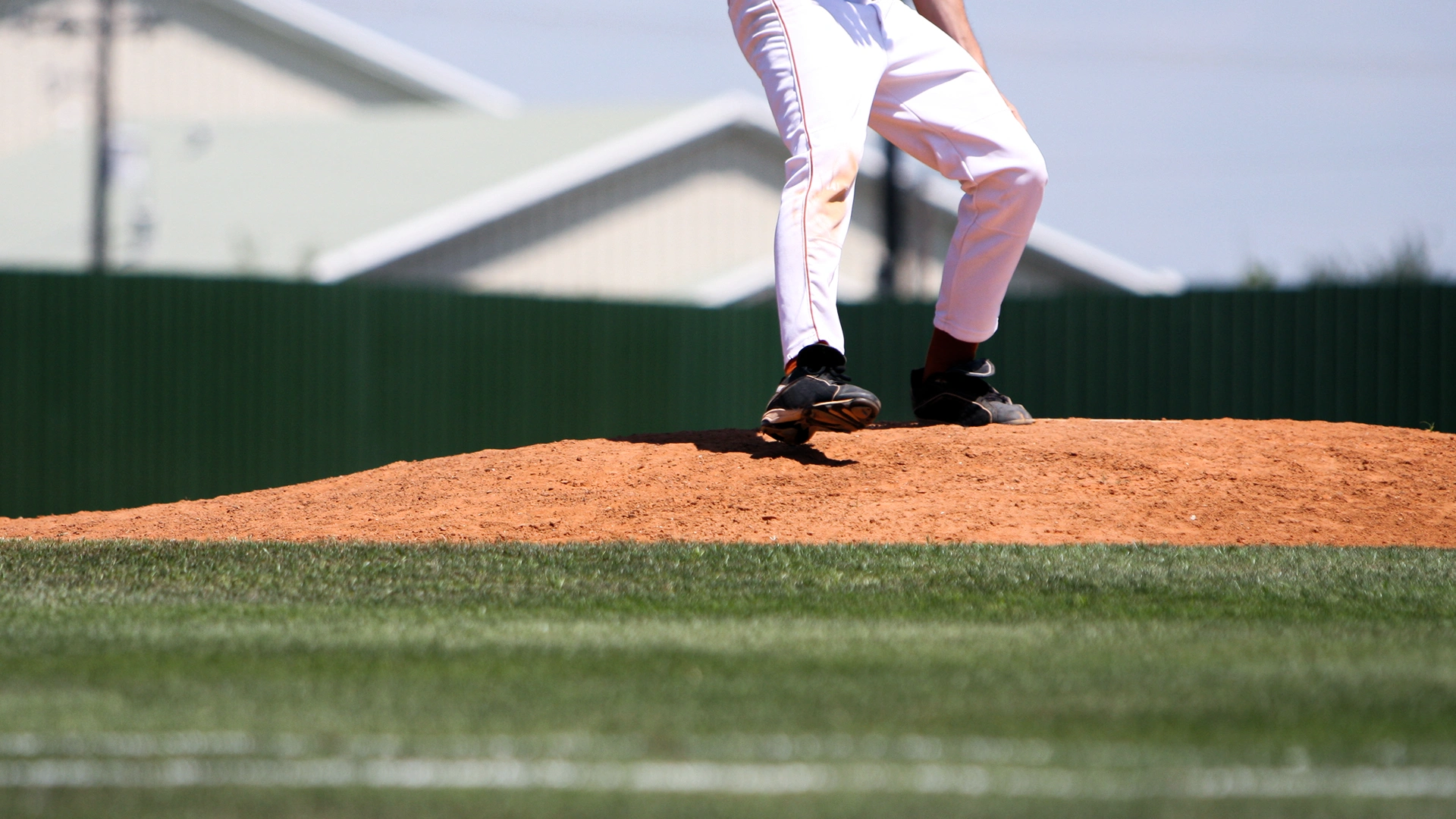 A baseball player stands on the pitchers mound, wearing white pants and black shoes, with buildings and a green fence in the background.