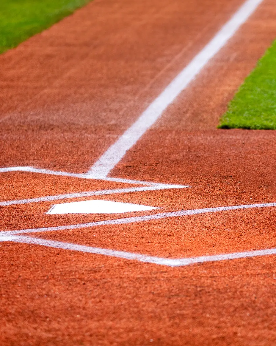 Close-up of a baseball home plate with white lines on a red clay field, bordered by green grass.