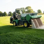 Groundskeeper driving a green utility vehicle spreading brown topdressing material onto a golf course fairway.