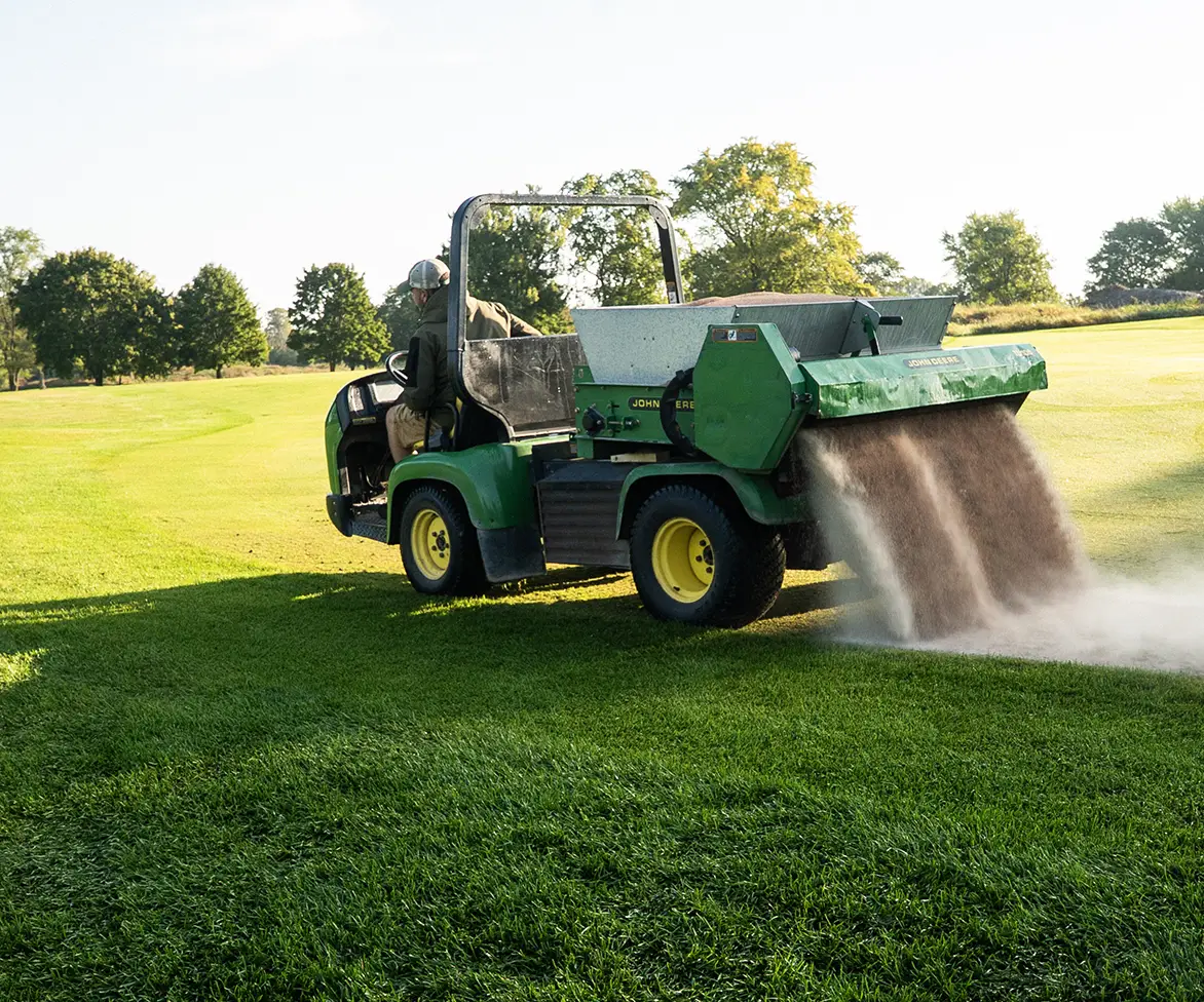 Groundskeeper driving a green utility vehicle spreading brown topdressing material onto a golf course fairway.