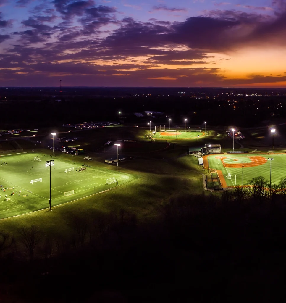 Aerial view of illuminated sports fields at dusk, with soccer and baseball fields lit up, surrounded by parking areas and distant city lights under a dramatic sunset sky.