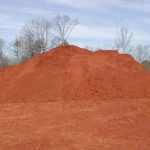 Large mound of bright red infield dirt piled outdoors, with bare trees and a cloudy sky in the background.