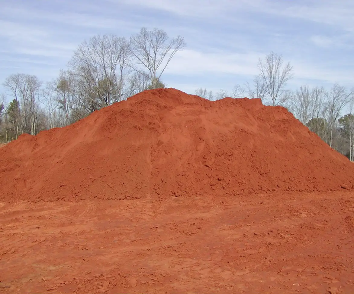 Large mound of bright red infield dirt piled outdoors, with bare trees and a cloudy sky in the background.