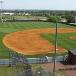 Elevated view of a baseball field with bright orange infield dirt and striped green outfield grass, seen from behind a fence with a few people standing near the dugout area.