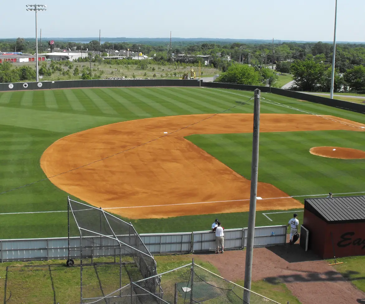 Elevated view of a baseball field with bright orange infield dirt and striped green outfield grass, seen from behind a fence with a few people standing near the dugout area.