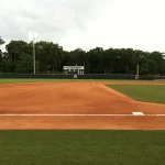 Baseball field with bright orange infield dirt and green grass outfield; a scoreboard in the distance reads “Atlantic Sun Championship,” with trees and stadium lights behind the outfield fence.