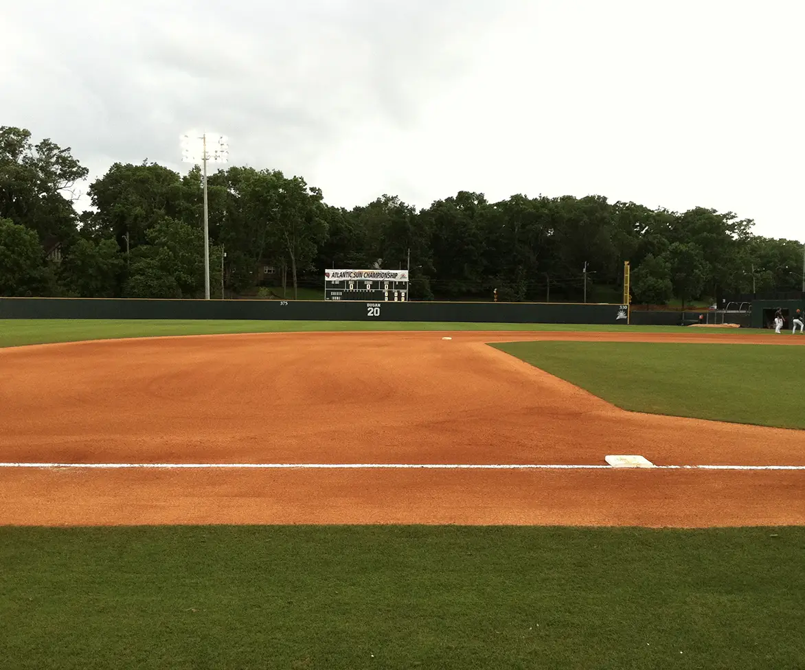 Baseball field with bright orange infield dirt and green grass outfield; a scoreboard in the distance reads “Atlantic Sun Championship,” with trees and stadium lights behind the outfield fence.