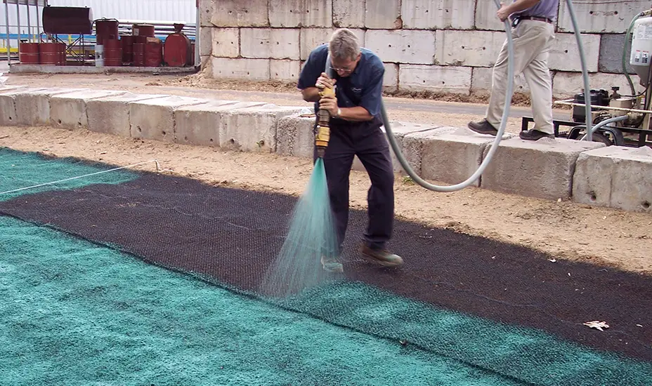 A worker sprays green hydroseed mulch onto the ground with a hose, while another person stands nearby. Concrete barriers and barrels are visible in the background.
