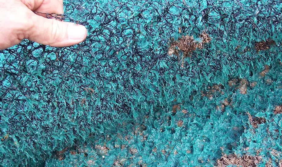 A hand lifts a thick, tangled green and black mesh mat, possibly used for erosion control or landscaping, showing its rough, fibrous texture and some dirt caught in the material.