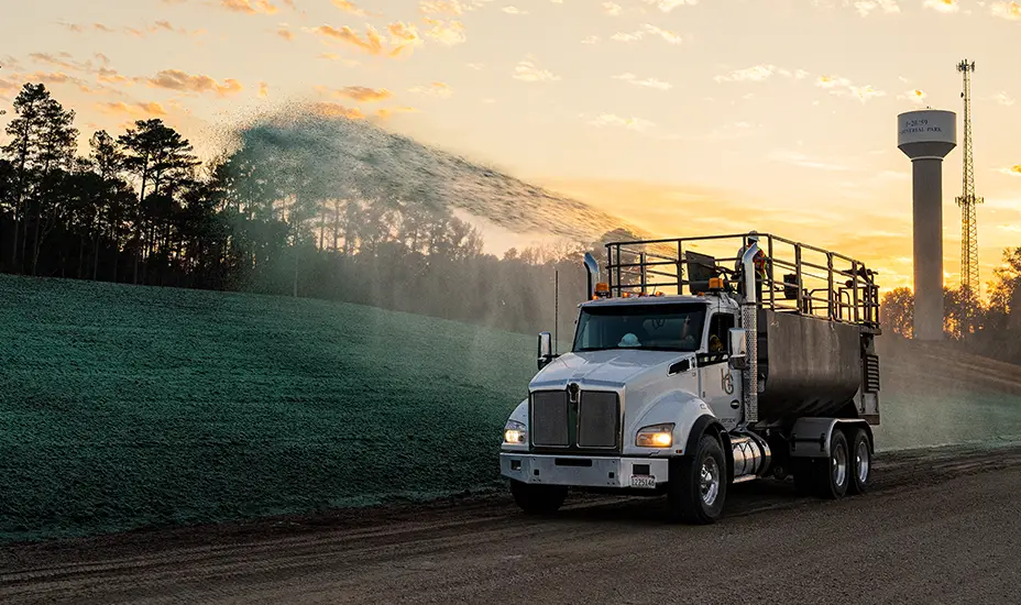 A truck sprays green material on a sloped roadside at sunset, likely for grass seeding or erosion control. Trees, a water tower, and a cell tower are in the background.