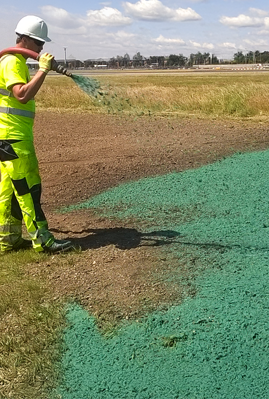 A worker in a safety vest and helmet sprays green hydroseed onto a bare patch of soil in an open field under a partly cloudy sky.