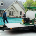 A person sprays green hydroseed onto a yard in front of a house, using equipment mounted on a trailer.