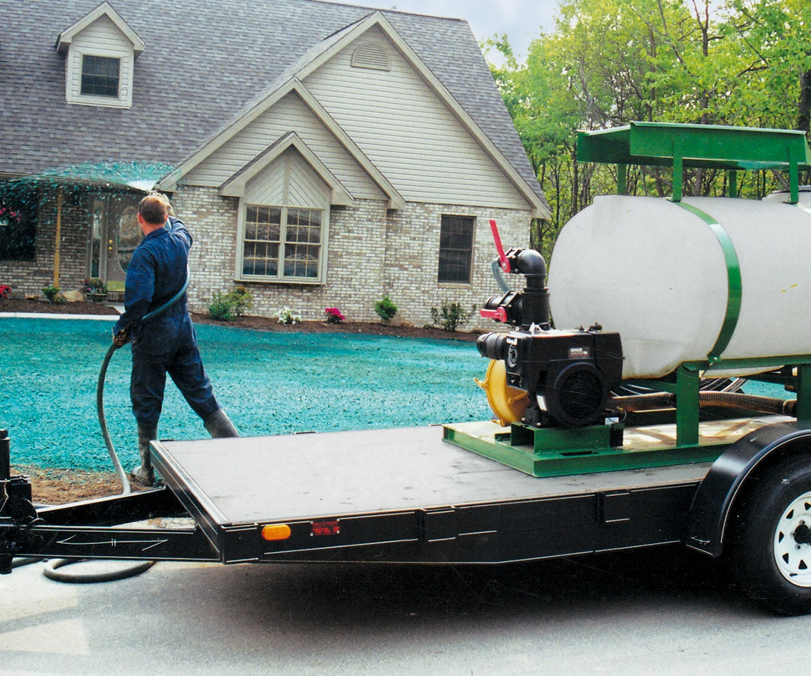 A person sprays green hydroseed onto a yard in front of a house, using equipment mounted on a trailer.