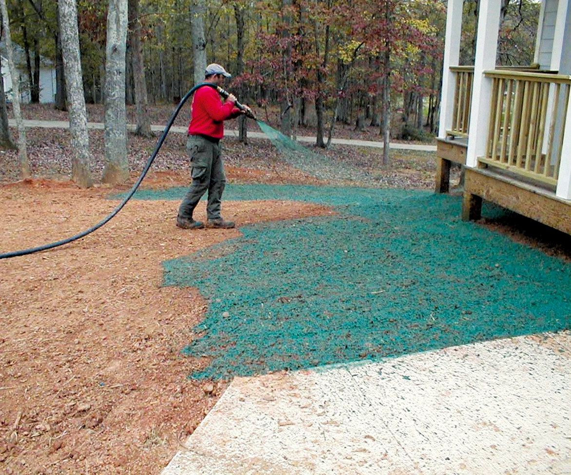 A person in a red shirt sprays green hydroseed over bare soil near a house porch, preparing the area for new grass growth. Trees with autumn leaves are in the background.