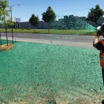 A worker in a safety vest sprays green hydroseed over bare ground beside a road with young trees, likely to promote grass growth.