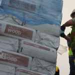 Construction workers in safety vests and helmets secure a large stack of packaged wood products wrapped in plastic. The packages are labeled WOOD and are being handled outdoors.