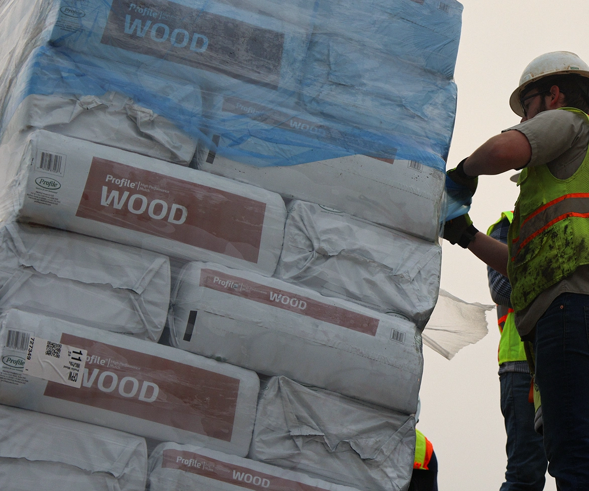 Construction workers in safety vests and helmets secure a large stack of packaged wood products wrapped in plastic. The packages are labeled WOOD and are being handled outdoors.