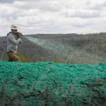 A person wearing a hard hat sprays green hydroseeding mixture onto a large, bare expanse of soil with trees and hills in the background under a cloudy sky.