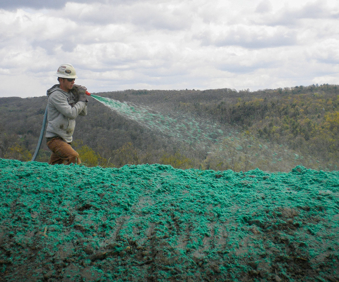 A person wearing a hard hat sprays green hydroseeding mixture onto a large, bare expanse of soil with trees and hills in the background under a cloudy sky.