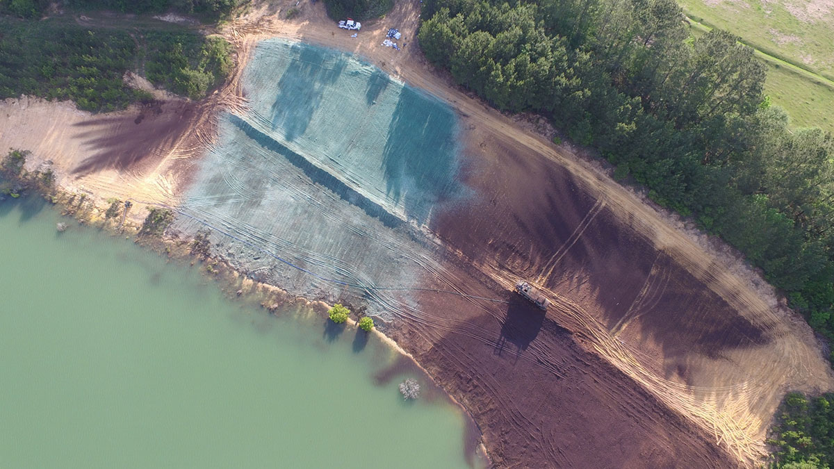 Aerial view of land by a body of water, showing areas with green and brown soil, construction equipment, and surrounding trees on one side.