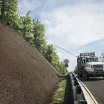 Hydroseeding truck spraying soil or mulch mixture onto a steep roadside embankment next to a highway guardrail.
