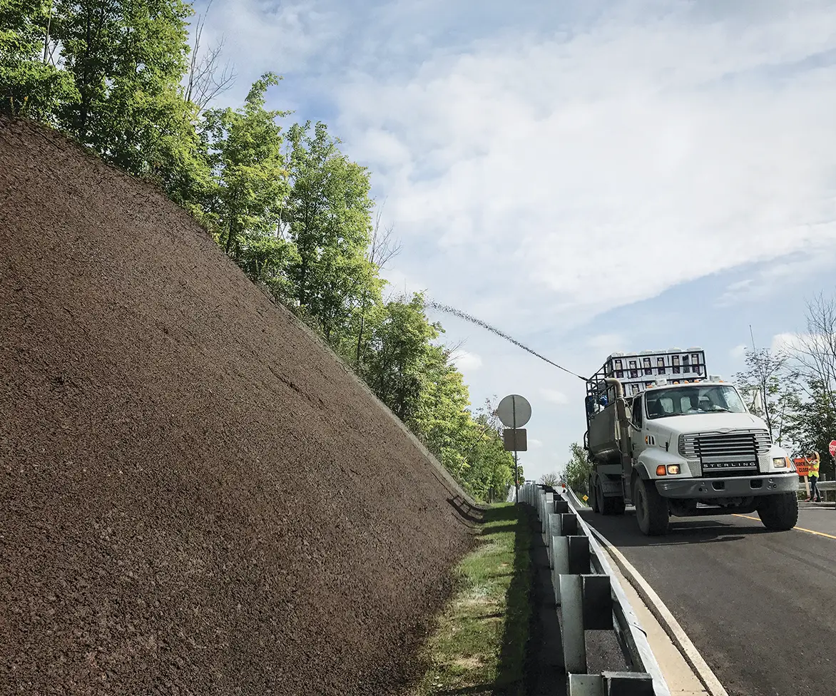 Hydroseeding truck spraying soil or mulch mixture onto a steep roadside embankment next to a highway guardrail.