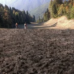 Wide view of freshly applied dark soil amendment across a cleared mountain roadway construction area with workers standing in the distance.