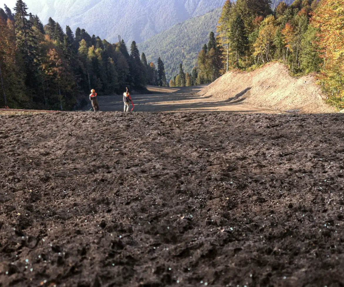 Wide view of freshly applied dark soil amendment across a cleared mountain roadway construction area with workers standing in the distance.