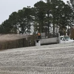 Worker operating a hydroseeding machine spraying a soil or mulch mixture onto a sloped construction site surrounded by trees.