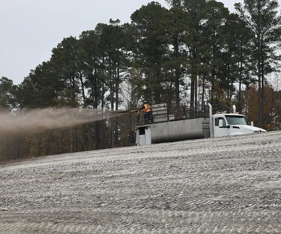 Worker operating a hydroseeding machine spraying a soil or mulch mixture onto a sloped construction site surrounded by trees.