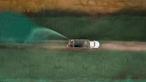 Aerial view of a white truck spraying water or liquid onto a dirt road, surrounded by green and brown agricultural or industrial fields.